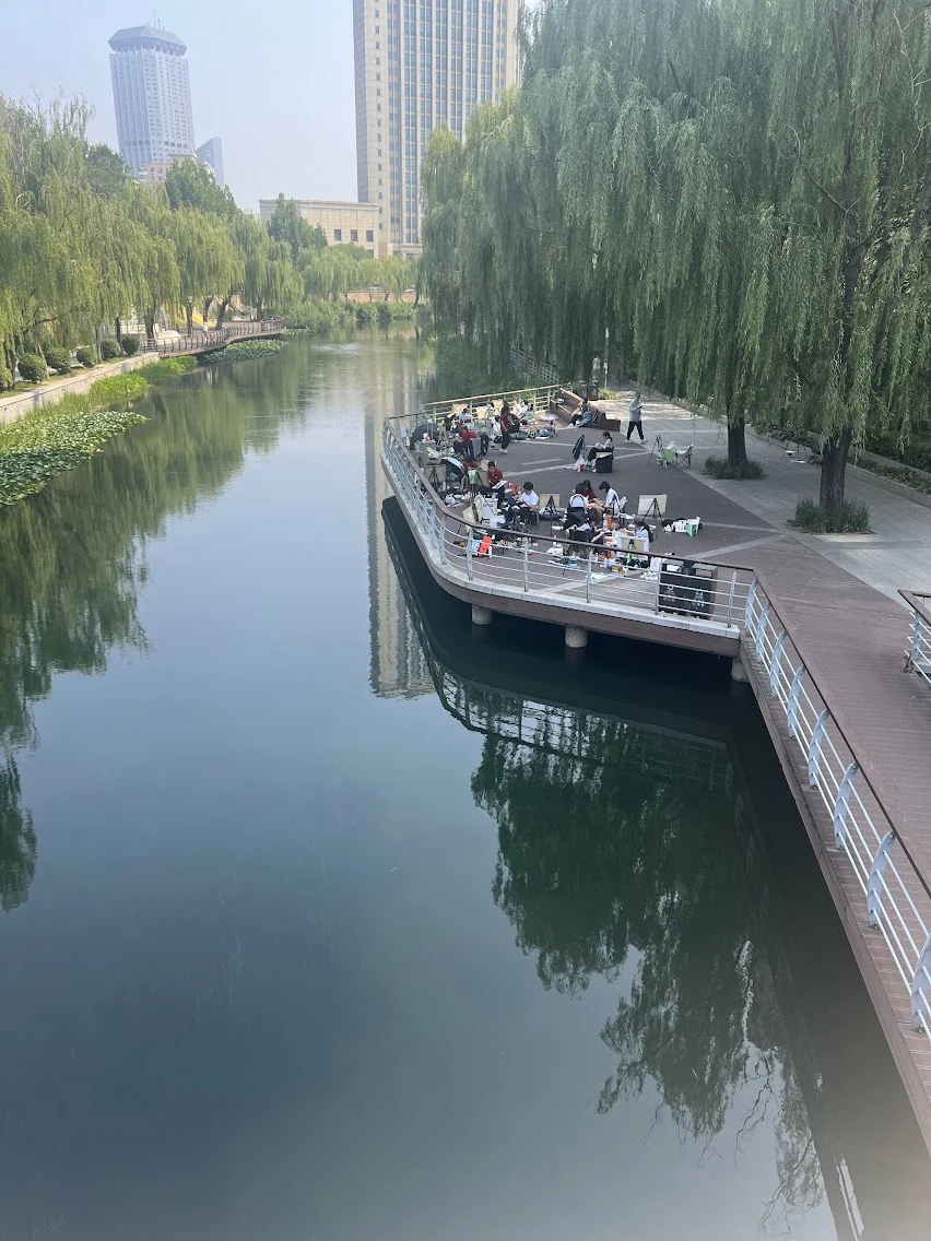 A tree-lined canal and riverside walkway, with people sitting along the water